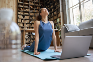 Mujer practicando yoga online desde casa frente a una laptop, realizando la postura de la cobra sobre colchoneta azul